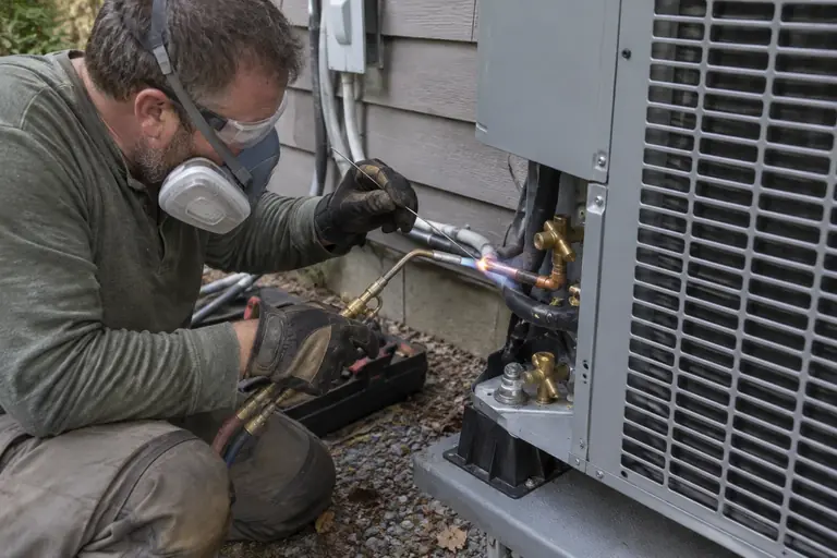 A technician in full protective gear is brazing and connecting a refrigerant line set to an outdoor heat pump unit beside a lap-sided home.