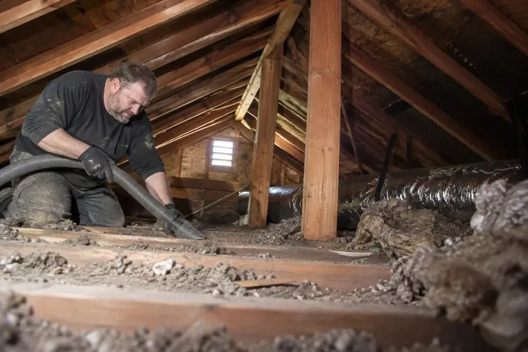 A technician uses a large vacuum hose to remove debris and old insulation from an attic in a Craftsman bungalow.