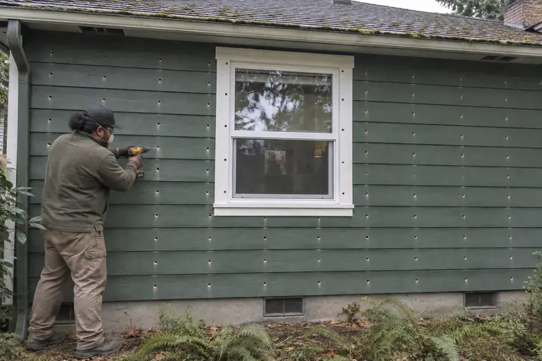 Exterior drill-and-fill on a Craftsman rambler, in progress: A technician drills evenly spaced holes through the green cedar siding to prepare for blowing in insulation into the empty wall cavities.