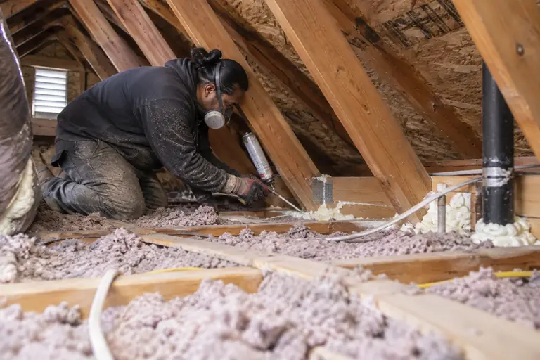 A technician air seals attic top plates and penetrations before adding fresh blown-in insulation in a Craftsman bungalow attic.