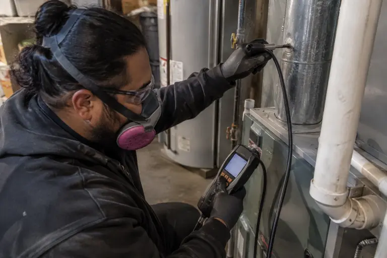 A technician in full protective gear is performing carbon monoxide and combustion analysis at the furnace flue inside a residential garage.