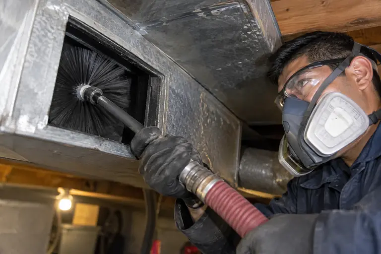 A technician fully geared in protective coveralls and safety equipment is using a rotating brush to agitate air ducts.