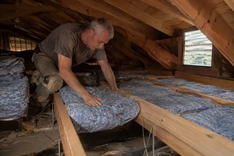 A technician carefully presses blue cotton denim batt insulation between crawl space floor joists supported by wire hangers.