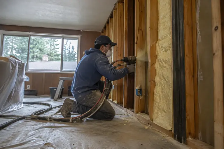 A technician in full protective gear applies closed-cell yellow-tan spray foam insulation into an open wall cavity between studs inside a mid-century rambler style home.