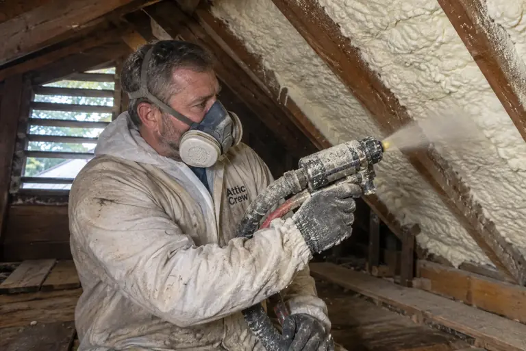 A technician in full protective gear sprays open-cell cream-white spray foam insulation on the underside of an attic roof deck between rafters.