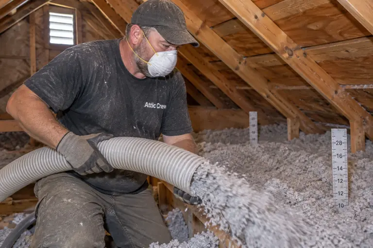 A technician installing loose-fill blown-in gray cellulose insulation across an attic floor with visible depth rulers in a Craftsman bungalow.