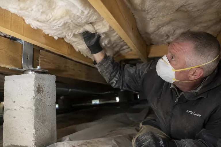 A technician installs under-floor insulation supported by metal braces in a crawl space beneath a raised foundation.