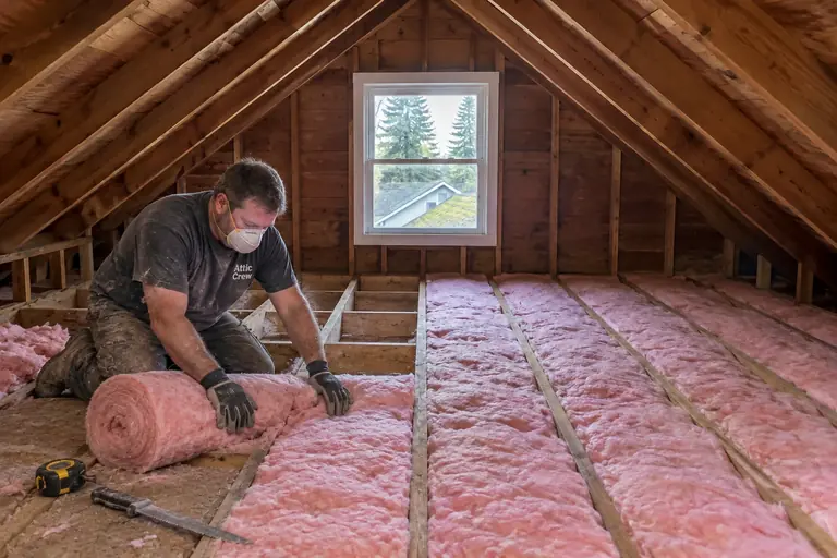 Attic insulation replacement in a Seattle Craftsman bungalow attic, in progress: Work is in progress with new pink fiberglass batts being unrolled and cut to fit snugly between the floor joists.