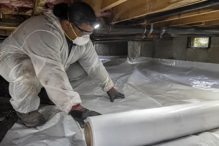 A technician carefully unrolls a heavy white 20-mil moisture barrier liner across the crawl space.
