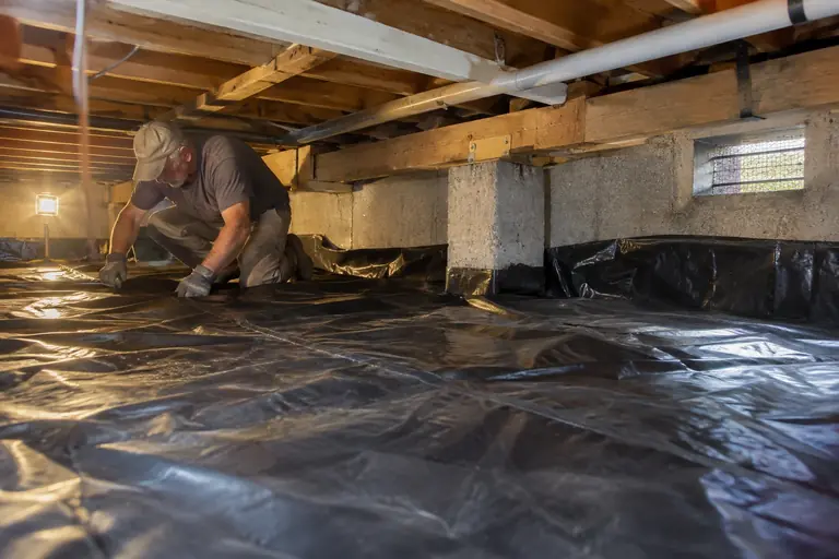 A technician installs a heavy white polyethylene vapor barrier over a damp dirt crawl space floor.