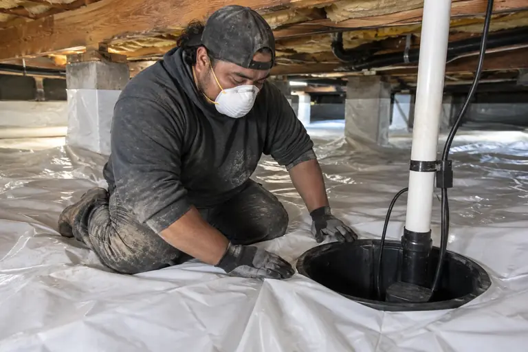Technician installs a sump pump basin beneath a freshly laid white moisture barrier liner in a damp crawl space.