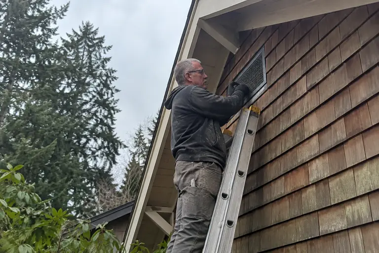 A technician is carefully screening a gable vent on a cedar-sided two-story home.